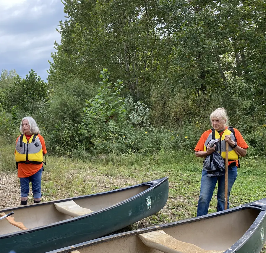 Two members in life jackets beside canoes on the bank of the Kokosing River
