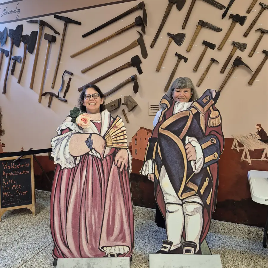 Two members posing in colonial cutout boards in front of a wall of antique tools